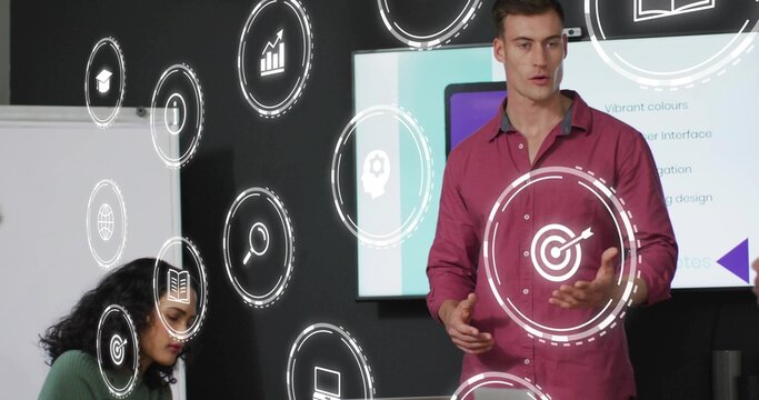 Presenting man wearing shirt gesturing at digital display in office, with laptop, icons, copy space - Powered by Adobe