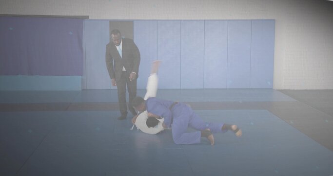 Blue gi fighter throwing white gi opponent on mats in training hall, coach in blazer observing