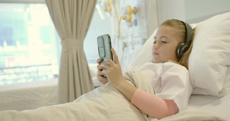 Lying child patient holding smartphone in hospital bed, wearing headphones near infusion pumps