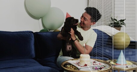 Adult man holding chocolate Labrador puppy at home party, with cake, pastel balloons, copy space