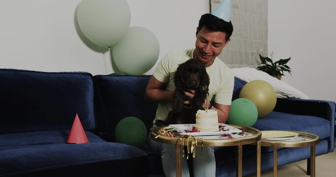 Celebrating man wearing T-shirt on blue sofa in living room, with puppy, cake and balloons - Powered by Adobe