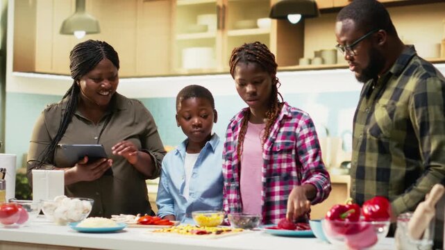 Smiling children following online recipe for pizza with their parents, preparing homemade food step by step with fresh ingredients. Black family gathered around the kitchen counter. Camera B.