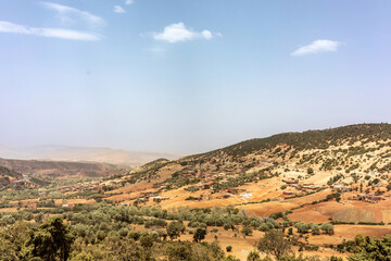 A vast semidesert valley between the Atlas Mountains and the Erg Chebbi dunes in Morocco, featuring dry hills, scattered acacia trees, and a partly cloudy sky.
