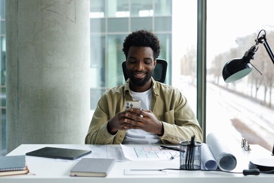 Front view of pleased young black male architect typing smartphone