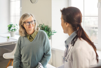 Positive elderly female patient consulting with a doctor or nurse in a hospital office during an...