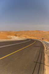 A curving desert highway with yellow road lines and a metal barrier disappears into a vast, golden expanse of sand dunes