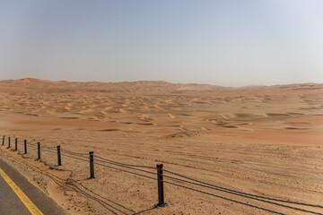 A desert highway curve with a simple wire fence overlooks a vast expanse of rippling golden sand dunes