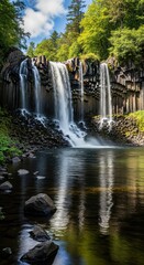 Multi-tiered Basalt Column Waterfall in Serene Setting. Reflective Pool with Scattered Foreground Rocks. Naturalistic Wilderness Cascade with Blue Sky.