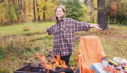 A beautiful woman stands joyfully by a campfire during a picnic in the woods. Dressed in a warm plaid shirt, she enjoys the vibrant autumn scenery filled with colorful leaves