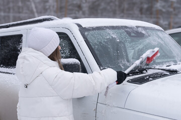A woman in a white jacket cleans snow and ice with a brush from the windshield of a white car in...
