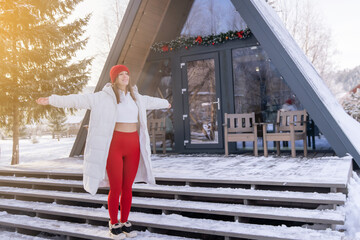 Young woman spreads her arms joyfully in front of modern triangular cabin surrounded by snow during winter morning light