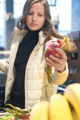 A young woman chooses fruits in a store, holding a red Pitahaya fruit in her hand. She emphasizes healthy eating while shopping with a basket.