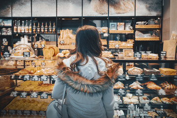 Woman examines and buys a display case with sweet cakes, croissants and buns. She wants to buy a sweet dessert for dinner, back view. Woman buys sweet desserts in bakery filled with cakes, croissants