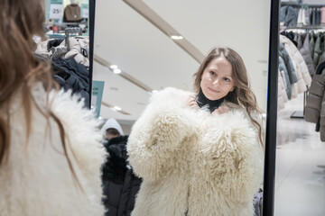 A white young beautiful 30-year-old woman tries on a white sheepskin coat in a clothing boutique, looking in the mirror. Seasonal fashion shopping and women's holiday shopping