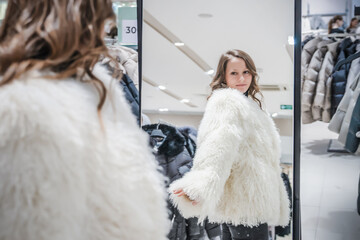 Young woman tries on a stylish white sheepskin coat in a clothing boutique while checking her reflection in the mirror during a seasonal shopping trip
