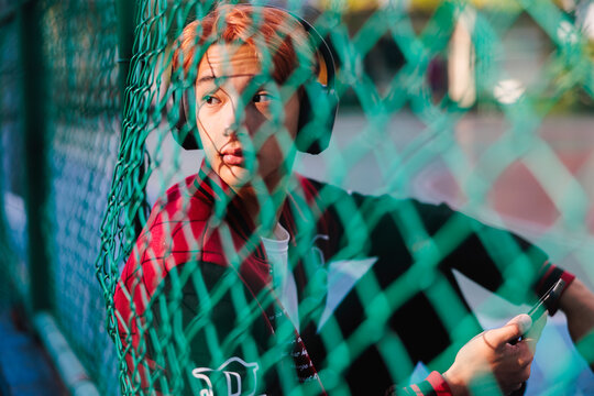 Man with headphones looks through fence, holding phone.
