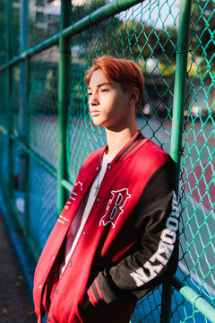 Man in varsity jacket leans against a chain-link fence.