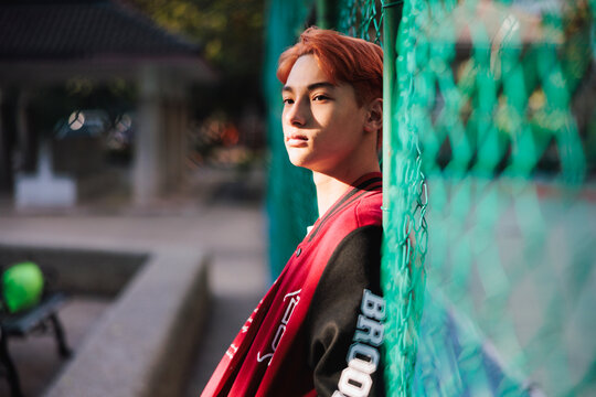 Pensive young man in varsity jacket by green fence.