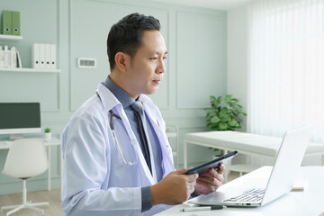 Portrait of asian senior doctor sitting in medical office while using laptop computer and checking time of appointment.