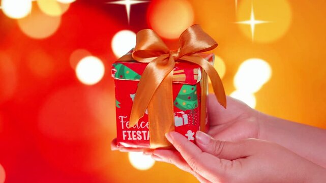 Hand holding Christmas gift with gold bow on red background with blurred lights