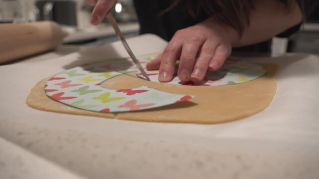 Unrecognizable woman cutting rolled dough with a knife using a paper template on kitchen counter. Homemade baking preparation, close-up cooking process.