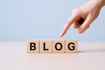 Blog Concept: Human Hand Pointing to Wooden Blocks Spelling BLOG on Table Against Blue Wall