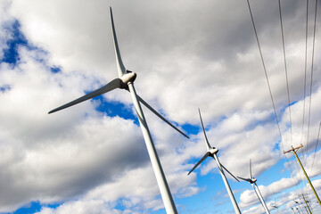 Windfarm with huge mills beside the power poles with electricity lines in the background of blue sky with white clouds.