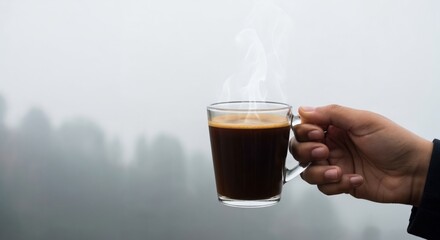 A person's hand gently holding a transparent glass cup filled with dark, steaming coffee, with a soft, misty forest in the background.