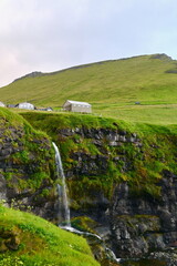Fototapeta premium Picturesque Village with Waterfall Flowing into the Ocean, Surrounded by Steep Cliffs and Green Hills in the Faroe Islands. Iconic Nordic Landscape