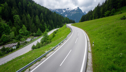 An empty, winding asphalt road curves through lush green meadows and dense forest in a valley, with snow-capped peaks in the background.