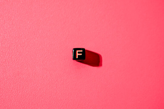 A stark, minimalist image featuring one black, square alphabet bead with the letter 'F' against a textured, vibrant pink background with dramatic shadow. - Powered by Adobe