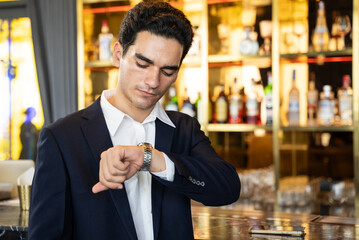 Young businessman checking his watch, standing at hotel bar counter. Concept of waiting, punctuality, after work networking, and luxurious evening lifestyle.