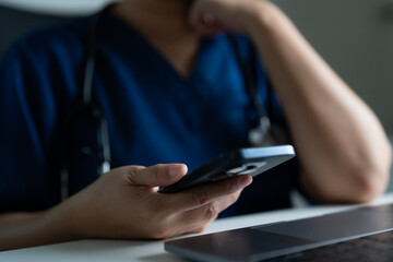 Doctor in blue scrubs using smartphone and laptop at desk, concept of telemedicine, online healthcare, digital consultation, and medical communication technology in modern hospital or clinic.