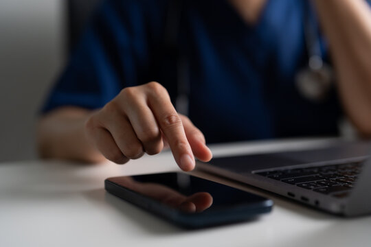 Doctor in blue scrubs touching smartphone next to laptop, concept of telemedicine, online healthcare service, telehealth technology, and digital medical communication in hospital or clinic office.