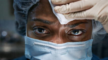 Close-up of a focused black female surgeon with worried look wearing surgical mask and cap, sweating and looking at camera with assistant wiping sweat from her forehead