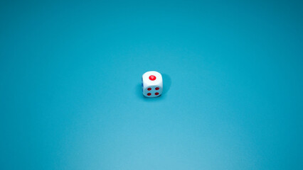An individual white game die is photographed at an angle on a solid teal blue background, showing...