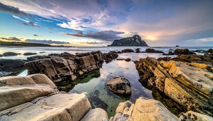 Rocky Shoreline With Footprints Leading To Calm Ocean Water Under A Dramatic Cloudy Sunset Sky With Distant Island Silhouette