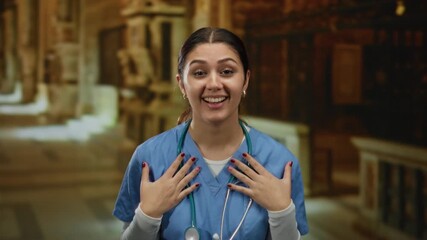 Young hispanic nurse in blue scrubs smiles in a church setting, conveying warmth and professionalism in an indoor environment.