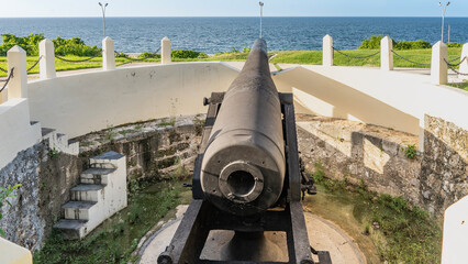 An antique firearm. The cannon is on a Taganana Hill, the barrel is pointing at the bay. The ocean is ahead. The fence is made of concrete pillars and chains. Battery of Santa Clara. Cuba. Havana.
