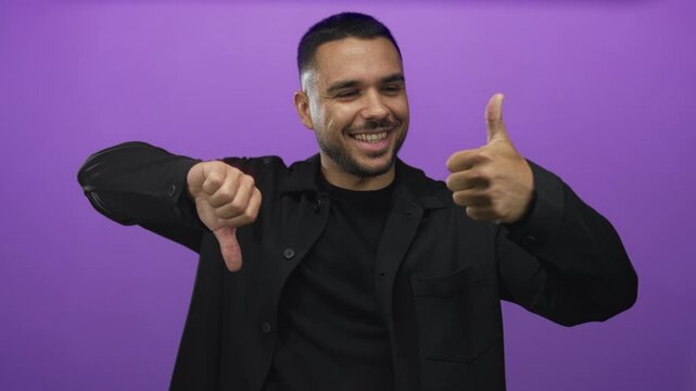 Young hispanic man wearing black shirt showing thumbs up and thumbs down gesture in purple studio setting; indecision.
