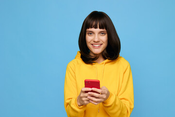 A cheerful young woman in a bright yellow hoodie uses a red smartphone against a solid blue backdrop, capturing a candid moment of everyday tech use and casual style. © SHOTPRIME STUDIO