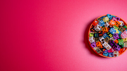 A vibrant assortment of multi-colored, square letter beads piled into an orange bowl, set against a bright, textured pink background.