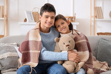 Happy brother and sister sitting on sofa in plaid with teddy bear in living room