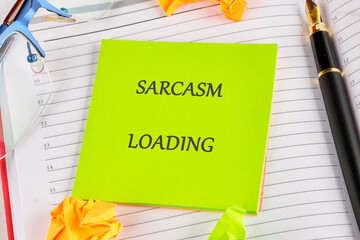 A bright green note with Sarcasm Loading is displayed on a planner next to a fountain pen, eyeglasses, and origami shapes. Be prepared for some sharp humor