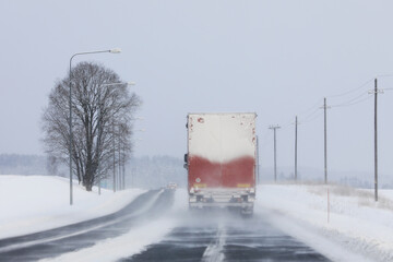 Road landscape of a semi truck driving along highway in snowfall on a day of winter. Difficult road conditions. Rear view, copy space.