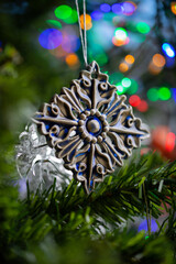 Ceramic toy in the form of a snowflake hanging on a Christmas tree with lights from a garland in the background.