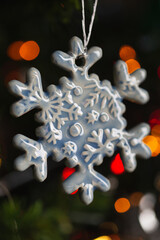 Ceramic toy in the form of a snowflake hanging on a Christmas tree with lights from a garland in the background.