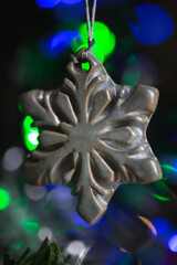 Ceramic toy in the form of a snowflake hanging on a Christmas tree with lights from a garland in the background.