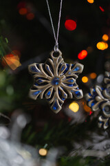 Ceramic toy in the form of a snowflake hanging on a Christmas tree with lights from a garland in the background.