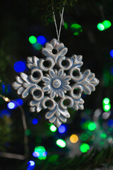 Ceramic toy in the form of a snowflake hanging on a Christmas tree with lights from a garland in the background.
